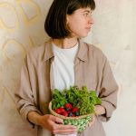 Woman standing with a basket of fresh greens and berries against a rustic wall background.
