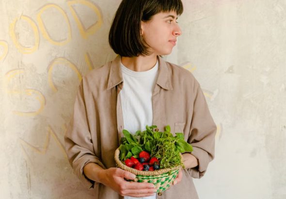 Woman standing with a basket of fresh greens and berries against a rustic wall background.